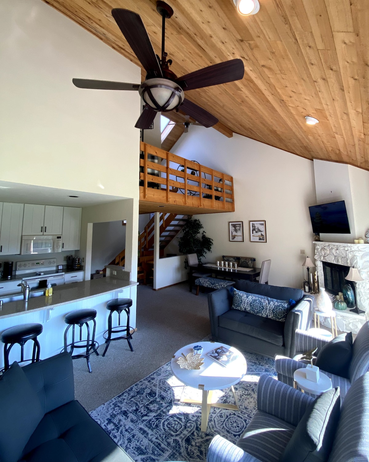 An open living area is highlighted by a vaulted ceiling with wooden beams. Comfortable seating surrounds a circular coffee table, with a fireplace visible in the background. A dining space and kitchen can be partially seen, featuring modern appliances and seating. A staircase leads to additional lofted accommodations.