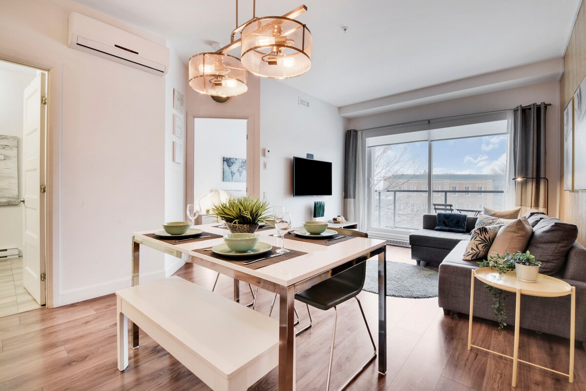 A modern dining area features a sleek table set for four, complemented by stylish chairs. A comfortable seating arrangement, with gray tones, is positioned nearby, and natural light flows through large windows, showcasing the view of a balcony with outdoor seating.