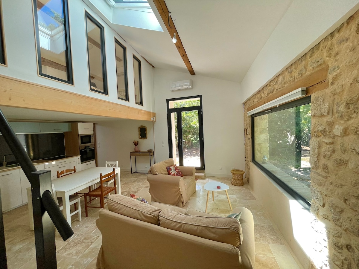 A spacious living area features a comfortable seating arrangement with a light-colored sofa. Large windows and a skylight illuminate the space, highlighting the stone wall and wooden beams. A dining table and chairs are visible, creating an inviting space that connects to the outdoors.
