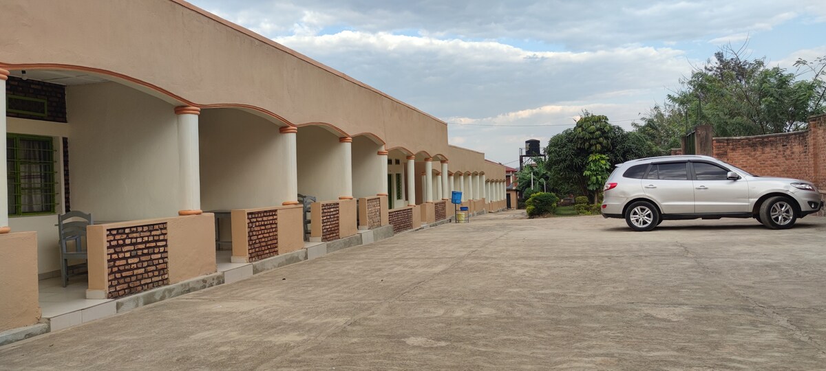 A view of the exterior of the motel features a row of arched doorways leading to individual rooms. A concrete driveway is visible, along with a parked SUV. Lush greenery surrounds the area, contributing to the serene environment.