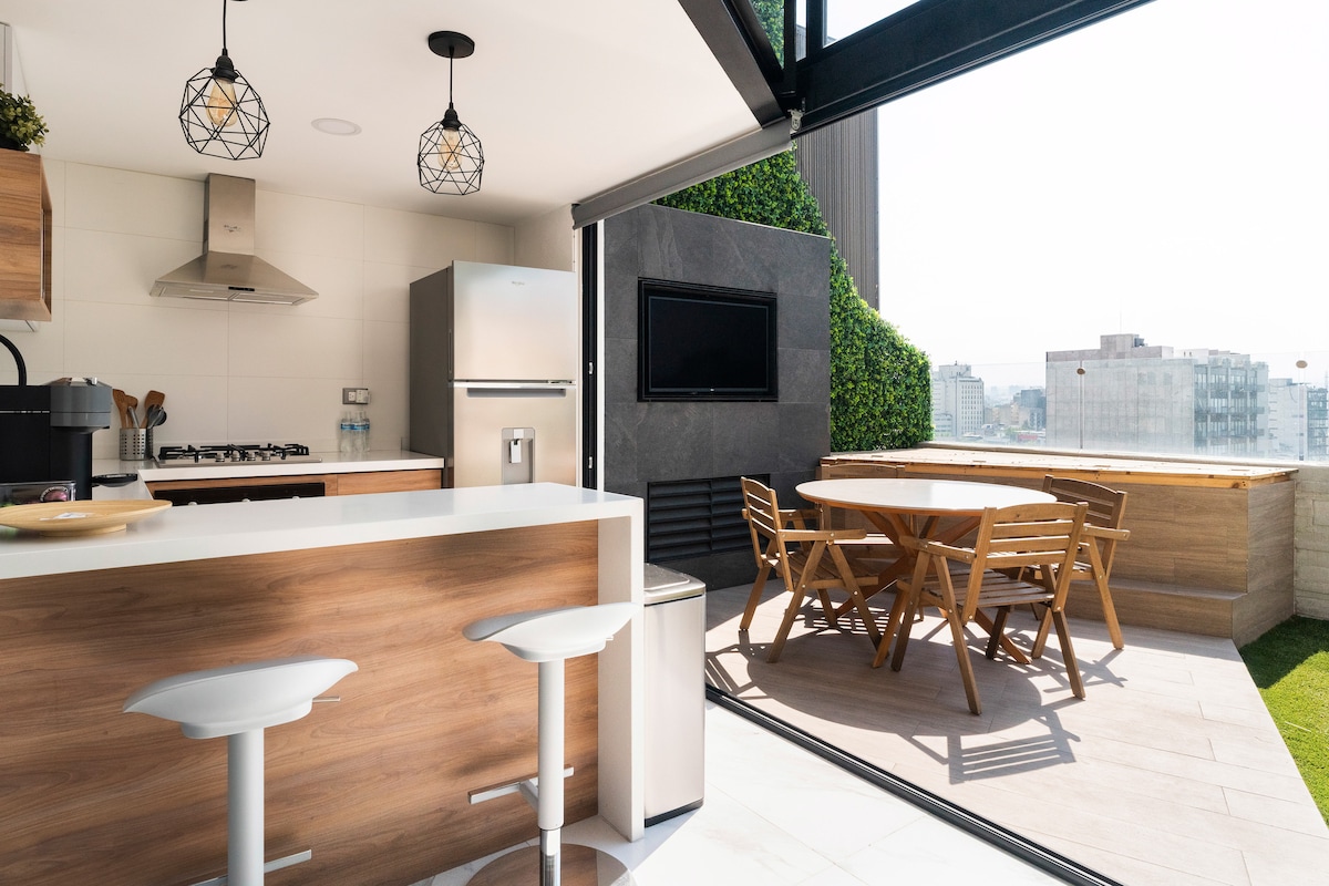 A modern kitchen features wooden cabinetry and stainless steel appliances, including a refrigerator and a stove. A dining area with a wooden table and chairs extends onto a terrace, where outdoor seating is arranged. Natural light fills the space, highlighting the green wall and city view.