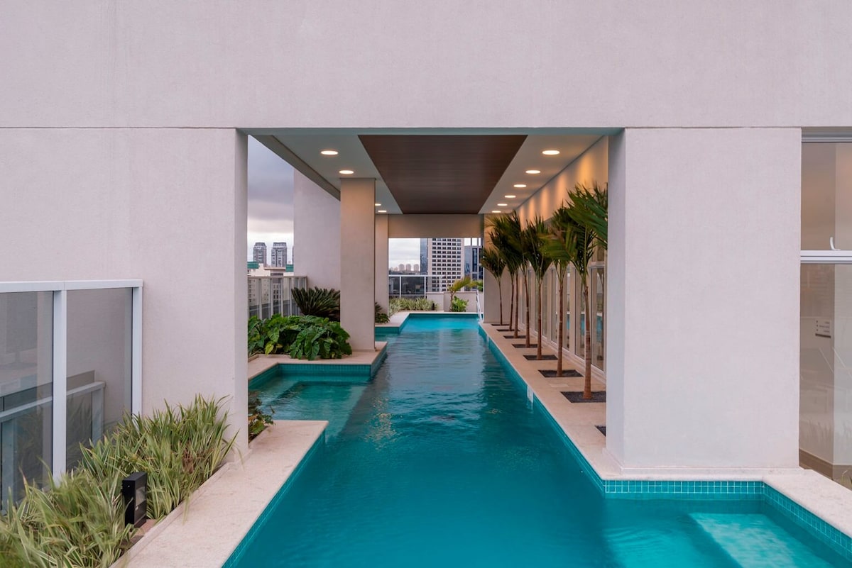A serene pool area is highlighted, featuring an elongated design with clear blue water. Lush greenery and palm trees are positioned along the edges. Natural light is provided through expansive windows, creating an environment that emphasizes tranquility and open space.