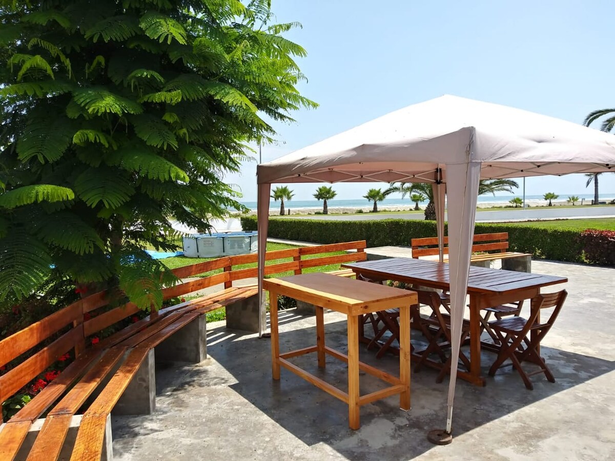 An outdoor seating area is presented under a canopy, featuring a wooden table and multiple benches arranged for gatherings. Lush greenery surrounds the space, complemented by a view of the water in the distance. The ground is paved, providing a clean and level surface.