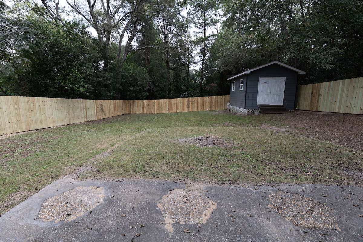 An outdoor area features a spacious grassy yard enclosed by a wooden fence. A small structure is visible in the background, complemented by a clear path leading to it. The surrounding greenery provides a natural backdrop, enhancing the area's openness and privacy.