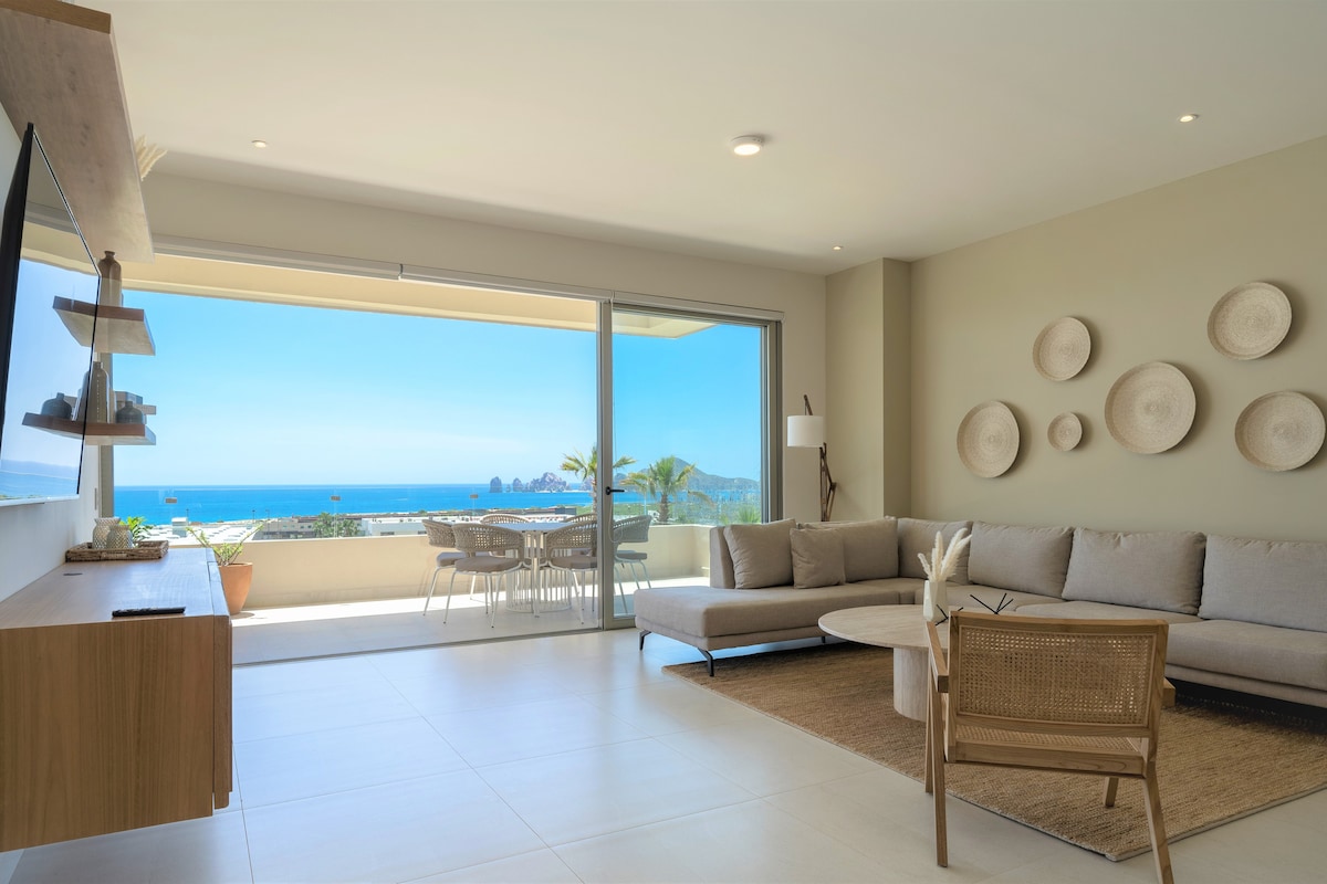 A modern living area features a spacious sofa arranged around a circular coffee table. Large glass doors offer an unobstructed view of the sea and a bright outdoor dining space. Natural light fills the room, highlighting the neutral color palette and decorative wall plates.