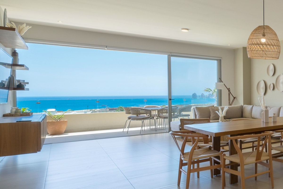 A spacious dining area features a wooden table surrounded by light-toned chairs. Large sliding glass doors provide a view of the ocean and a distant rock formation, while natural light fills the space, highlighting the light-coloured walls and minimalist decor.