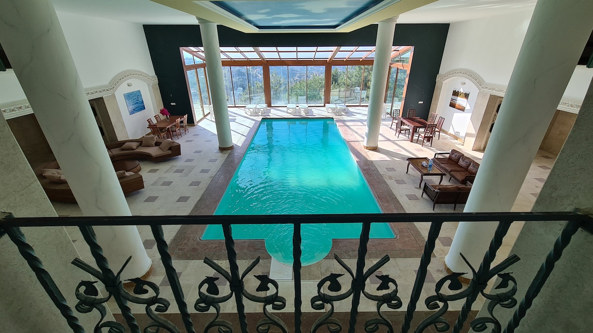 An indoor swimming pool is highlighted with crystal-clear water, framed by large glass windows that offer views of the surrounding hills. Comfortable seating areas and a dining table are positioned nearby, creating an inviting space for relaxation and gathering.