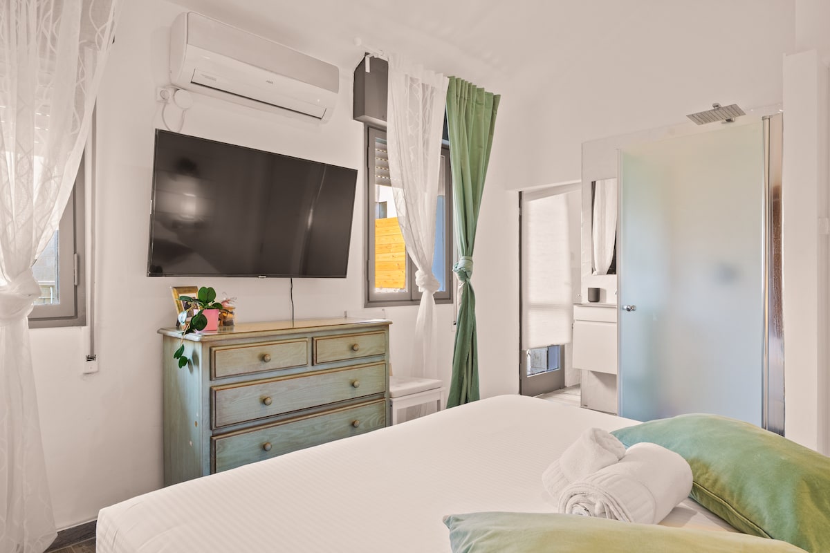The room features a spacious bed adorned with green accent pillows. A television is mounted on the wall opposite, with a light wooden dresser nearby. Sheer curtains frame the windows, allowing natural light to fill the space, while air conditioning is visible for comfort.