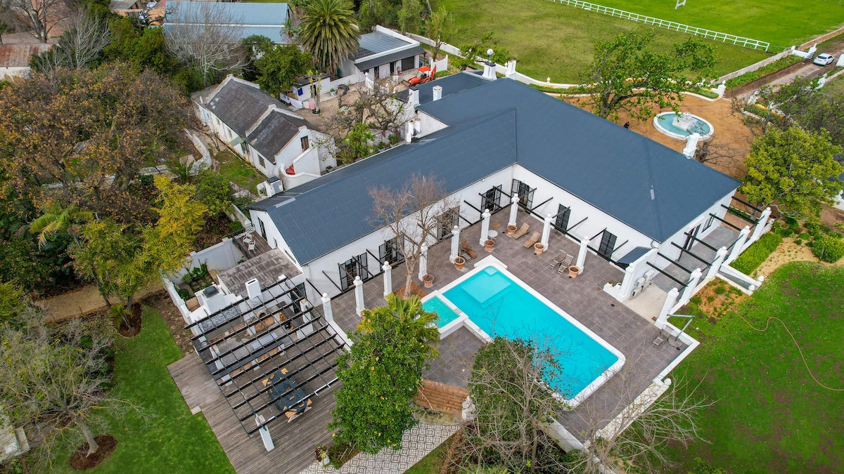 An aerial view presents the spacious Broadlands Manor, featuring a central pool surrounded by sun loungers. Lush greenery and landscaped gardens enhance the outdoor areas, while the building's sleek roof directs attention towards the inviting courtyard and various relaxation spots.