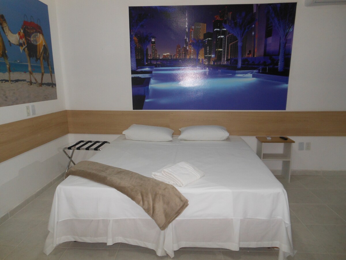 A spacious bed is positioned against a large mural featuring a city skyline at night. Soft linens and a beige throw blanket are neatly arranged over the bed. A small side table and a folding stool are visible in the room, enhancing functionality.