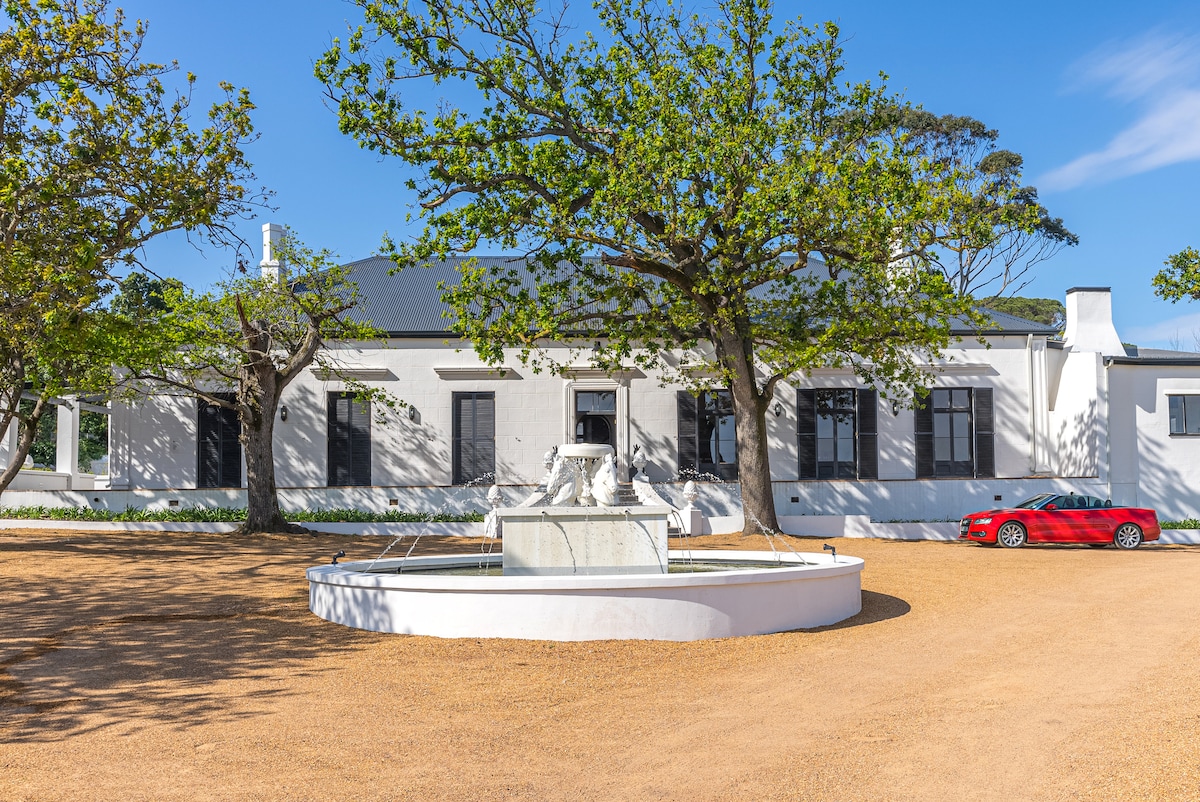 An elegant historic manor is presented with a circular fountain at its center, surrounded by well-maintained grounds. Large windows frame the facade, enhancing the architectural details. A red vehicle is parked nearby, providing a pop of color against the earthy gravel driveway.