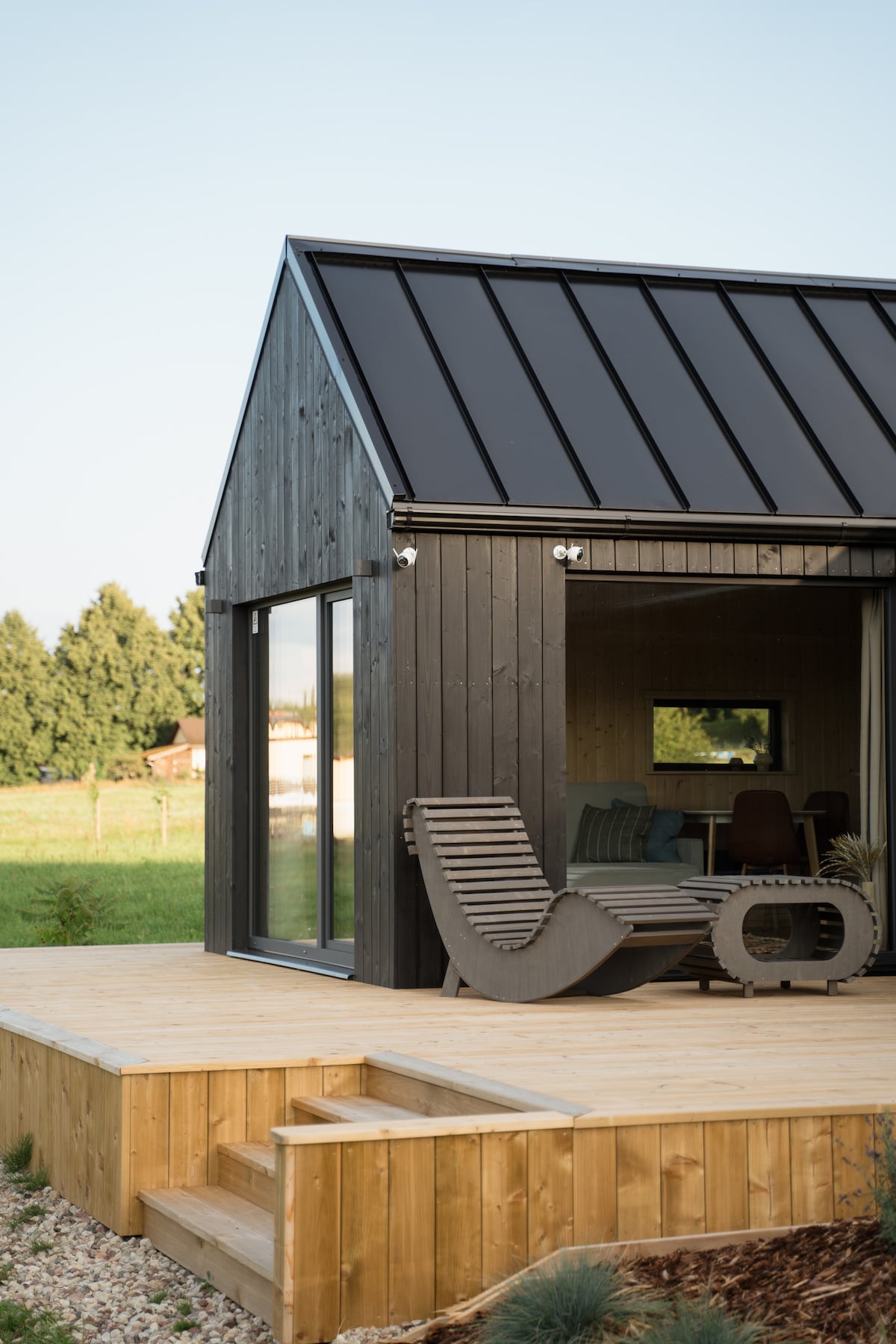 A modern cabin features a dark wood exterior with a peaked roof. Large glass windows provide views of the surrounding greenery. A comfortable lounge chair rests on a wooden deck, which is complemented by steps leading down to a landscaped area.