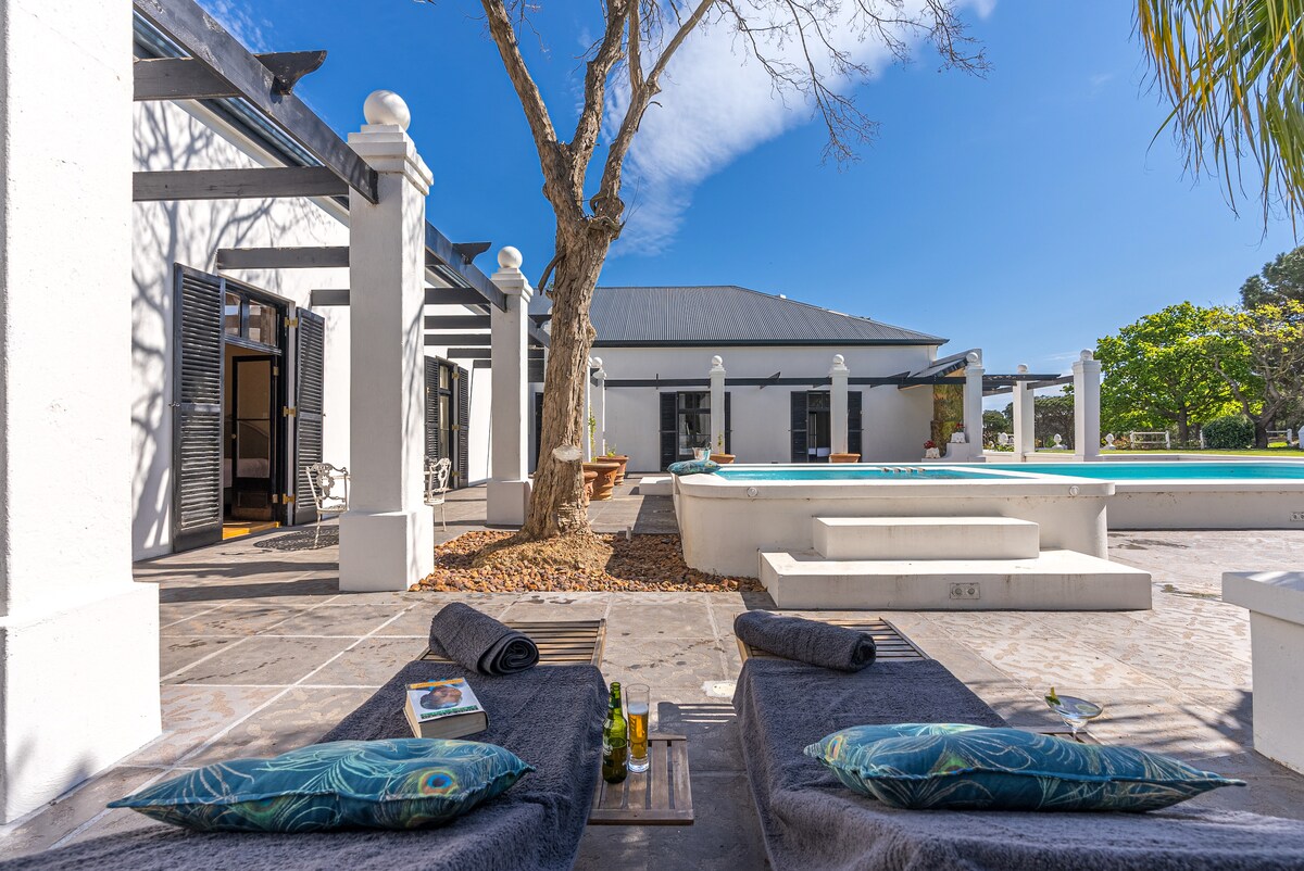Lounge chairs are positioned around a tranquil outdoor area, featuring a clear blue pool and surrounding greenery. A tree provides partial shade, while the bright sky adds to the serene atmosphere. Decorative elements, including a book and drinks, enhance the inviting space.