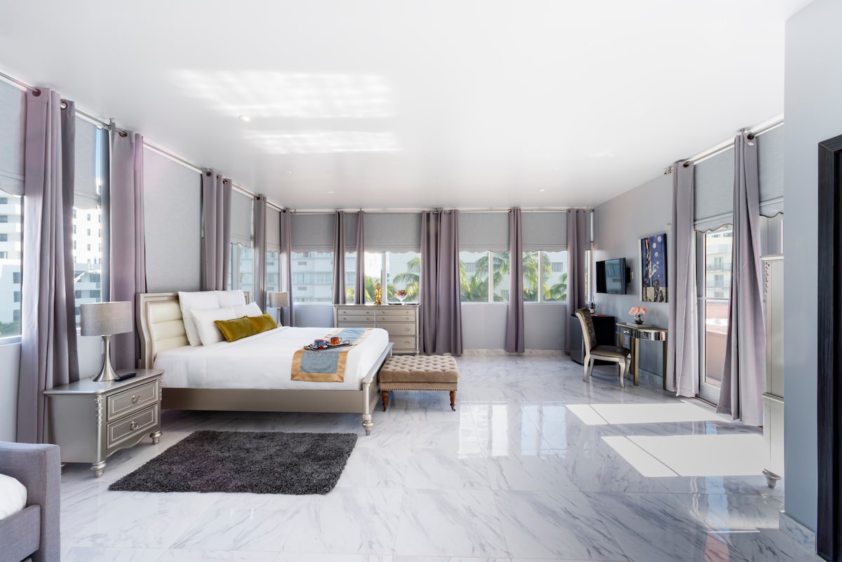 A spacious bedroom features a large bed with neutral bedding, enhanced by a decorative throw. Natural light floods through multiple windows, complemented by soft grey curtains. A plush area rug lies on the polished floor, while a seating area is furnished with a stylish chair and a small table.