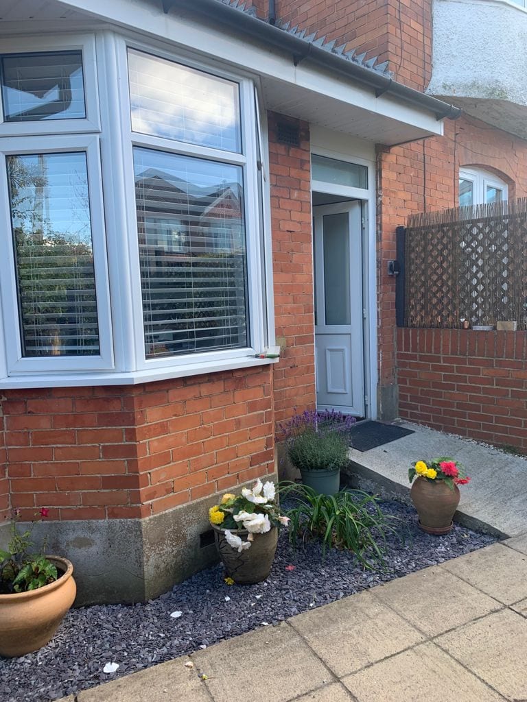 A welcoming entrance is framed by a large bay window with slatted blinds. Potted flowers and plants add a touch of greenery to the brick exterior, while a stone pathway leads to the front door, which is adjacent to a wooden lattice gate.