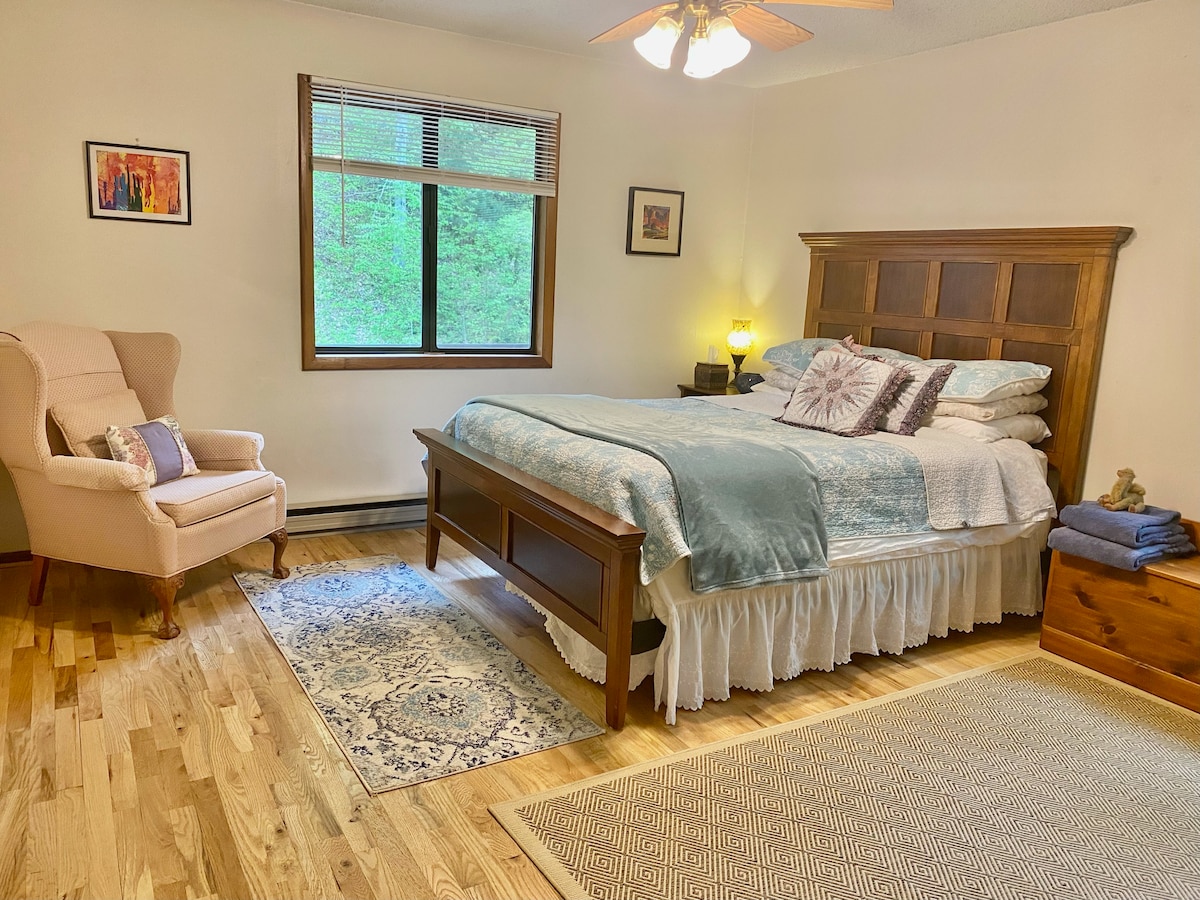 A spacious bedroom is furnished with a wooden bed dressed in soft linens and a decorative throw. A cozy armchair in the corner complements a bedside lamp, while large windows invite natural light and views of the surrounding greenery.