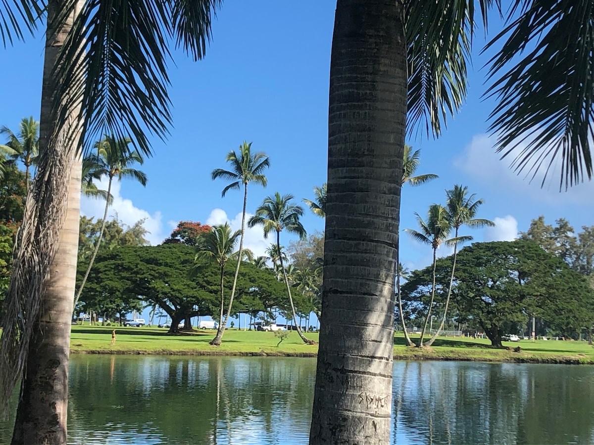 Lush palm trees frame a serene view of a calm waterway, reflecting the blue sky and scattered clouds above. In the background, an expanse of green lawn leads to tall trees and hints of coastal scenery, creating a peaceful outdoor atmosphere.