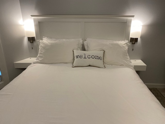 A spacious bed is centered in the image, featuring white bedding and two decorative pillows. A small pillow with the word 'welcome' is placed in the middle. Soft wall sconces are mounted on either side of the headboard, providing gentle lighting.