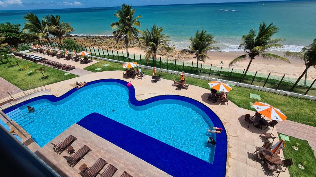A large swimming pool is set against a backdrop of the beach and ocean. Lounge chairs with umbrellas are arranged around the pool area, surrounded by lush palm trees and well-maintained greenery. The calm waves of the sea are visible in the distance.