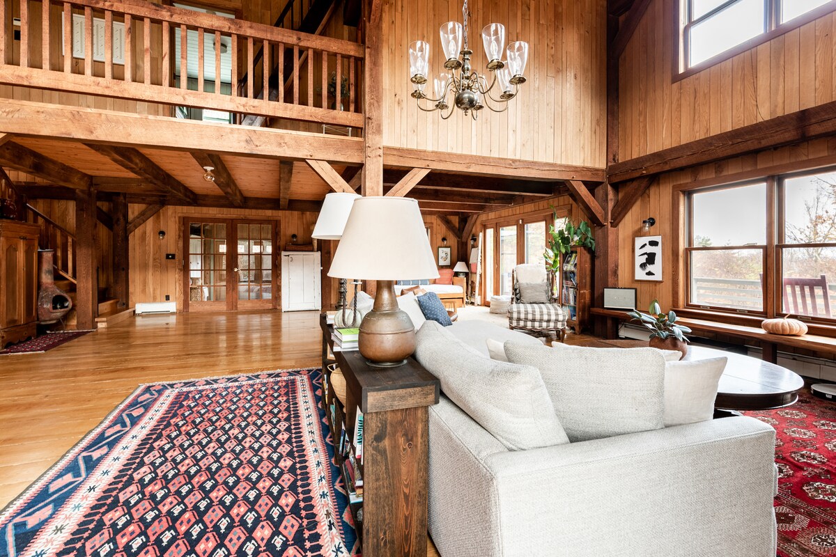 The spacious living area features wooden beams and warm-toned walls, creating a rustic ambiance. A comfortable sofa is positioned near a large window, where natural light floods the room. A stylish chandelier hangs above, complemented by a patterned area rug on the hardwood floor.