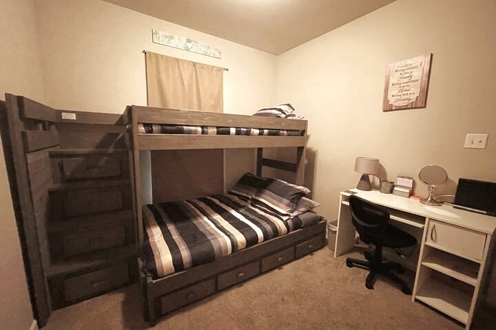 A multi-functional bedroom is furnished with a bunk bed featuring a double on the bottom and a twin on top. A comfortable desk with a chair is positioned nearby. Soft carpet complements the neutral wall color, creating a cozy environment.