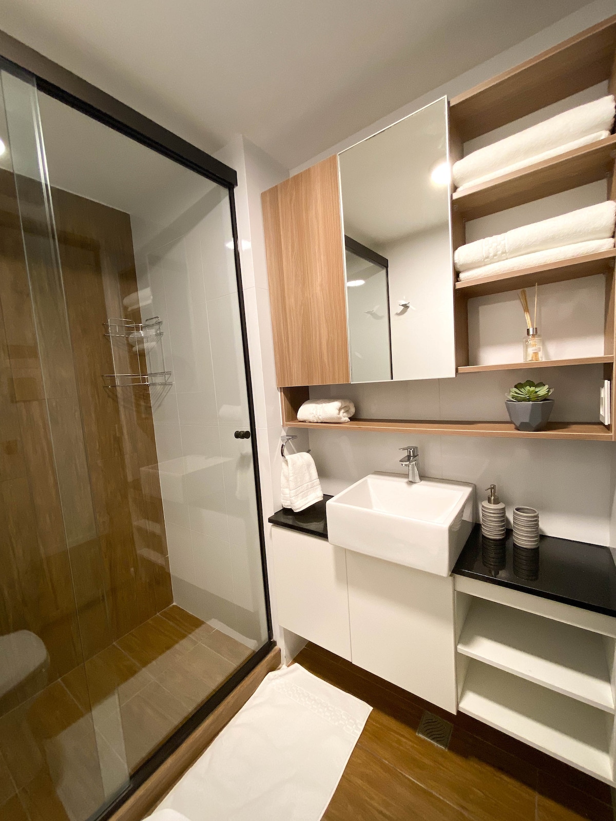 A modern bathroom with a glass shower stall and wooden paneling features a sleek sink and ample storage space. White towels are neatly arranged on shelves, and a small potted plant adds a touch of greenery. Natural light illuminates the space through a window.