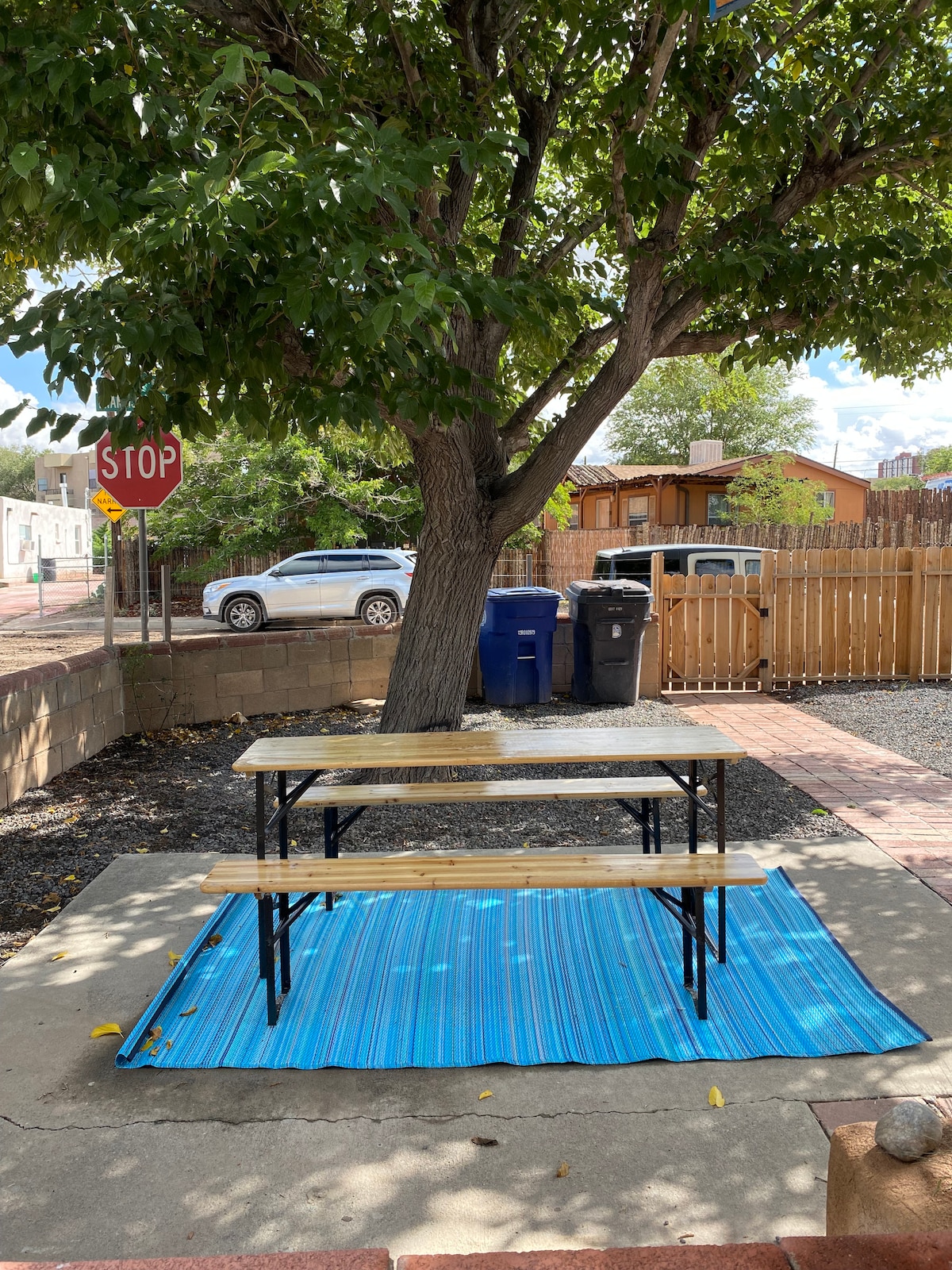 An outdoor dining area is visible, featuring a wooden table with attached benches. Beneath the shade of a tree, a blue rug adds color to the concrete surface. Nearby, a stop sign and a few trash bins are positioned, with a wooden fence in the background.