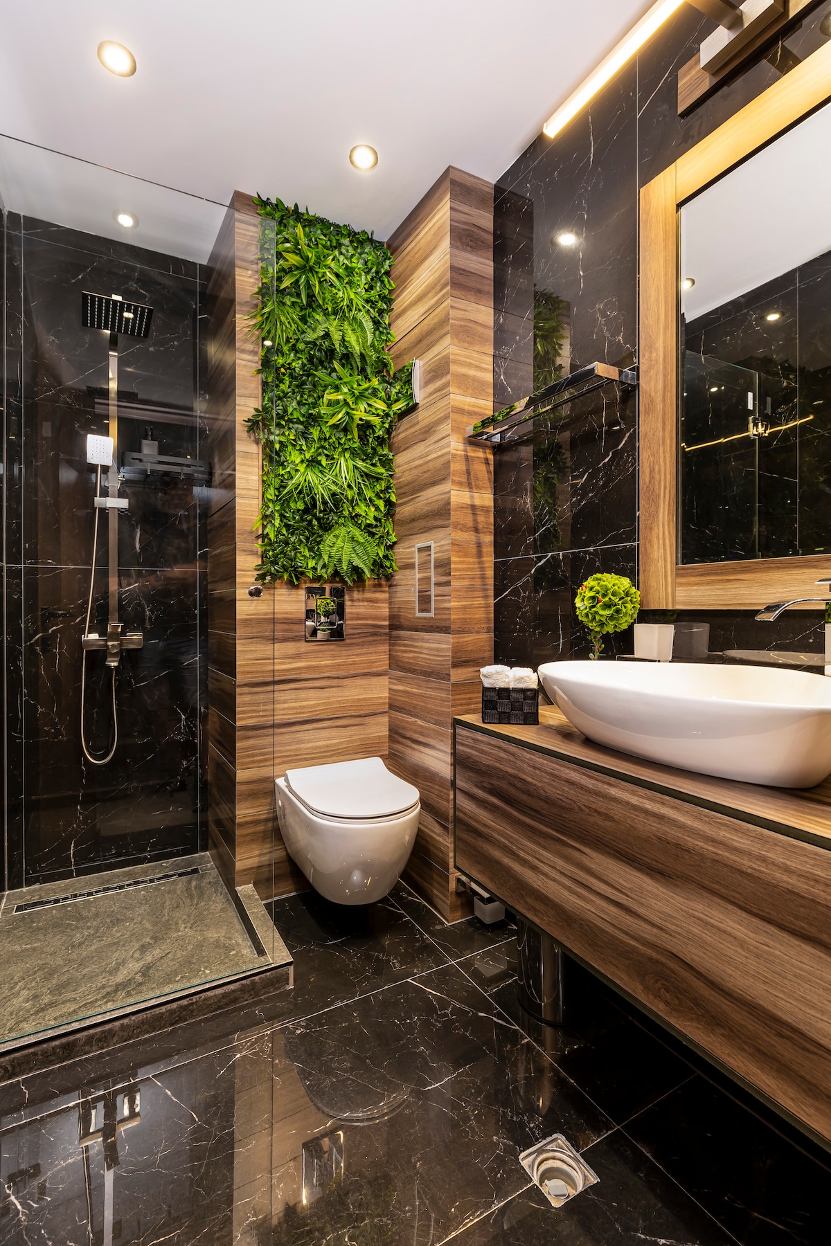 A modern bathroom features dark tile walls and stylish wooden accents. A walk-in shower with a rainfall showerhead is complemented by a vertical garden element. A sleek sink is positioned on a wooden vanity, with a large mirror reflecting the contemporary design.