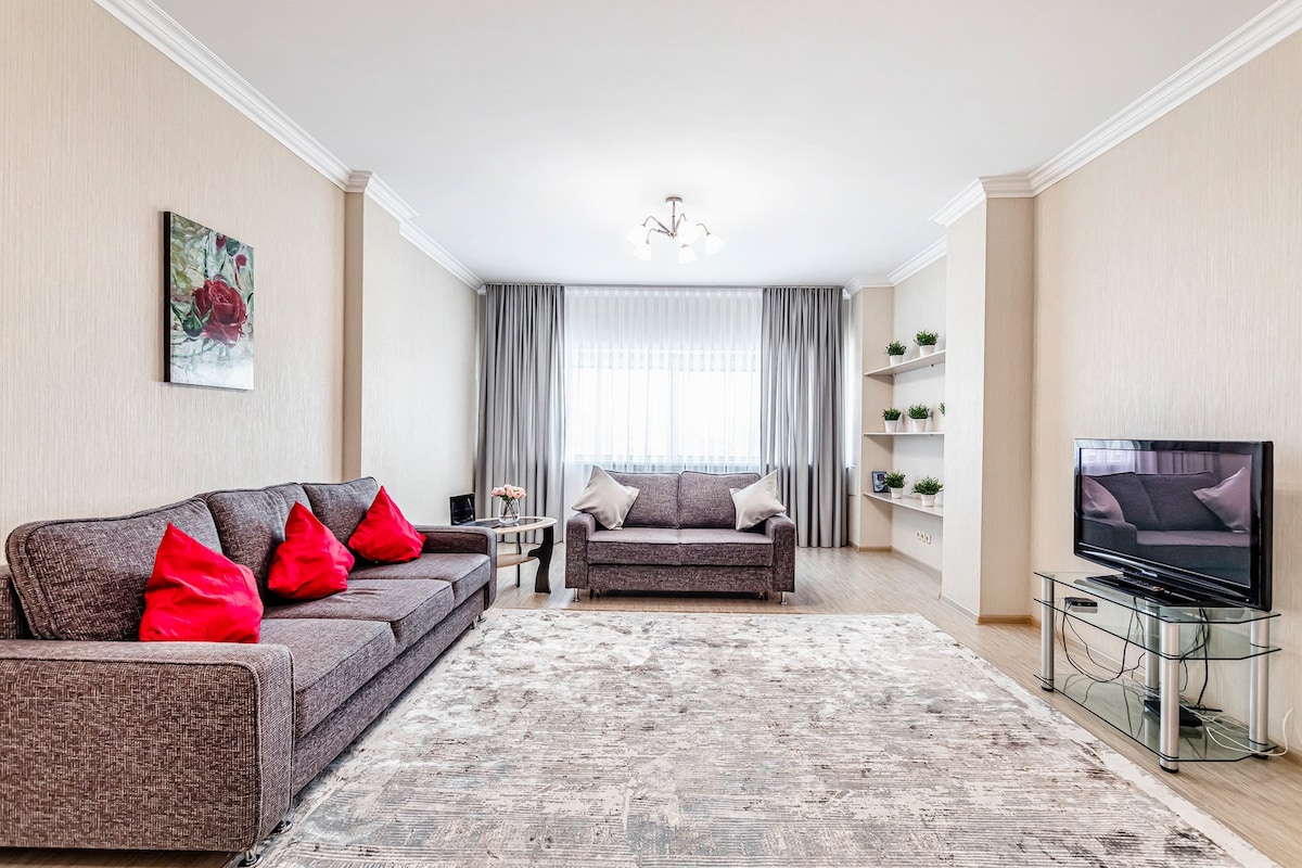 A spacious living area is presented, featuring two grey sofas adorned with bright red cushions. A television sits on a glass stand across from the seating arrangement. A soft, light-colored area rug covers the floor, and natural light filters through sheer curtains on the large window.
