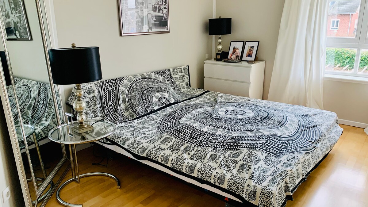 A spacious bed is adorned with a black and white patterned cover, set against neutral-colored walls. A round side table with a modern lamp is positioned next to the bed. Framed photographs are displayed on the surface, and a large mirror reflects the inviting room.