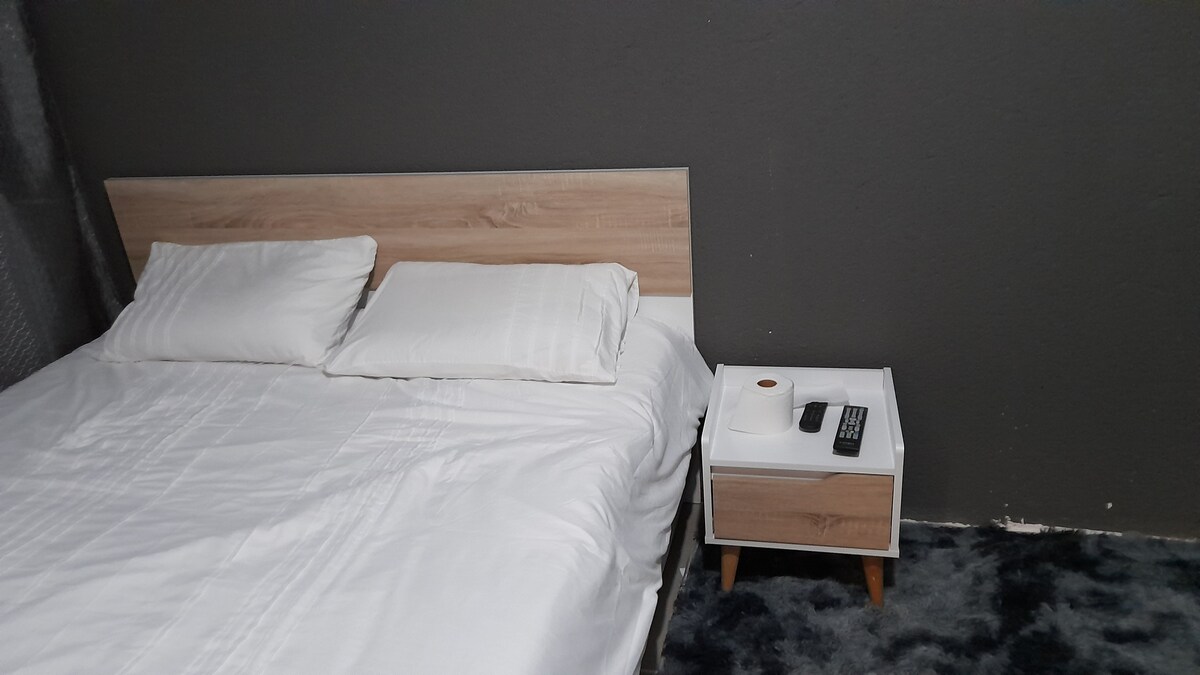 A cozy sleeping area features a wooden bed frame with a spacious bed dressed in white linens. A modern bedside table is positioned next to the bed, holding a remote and a small round object. Dark grey walls complement the warm tones of the furniture.