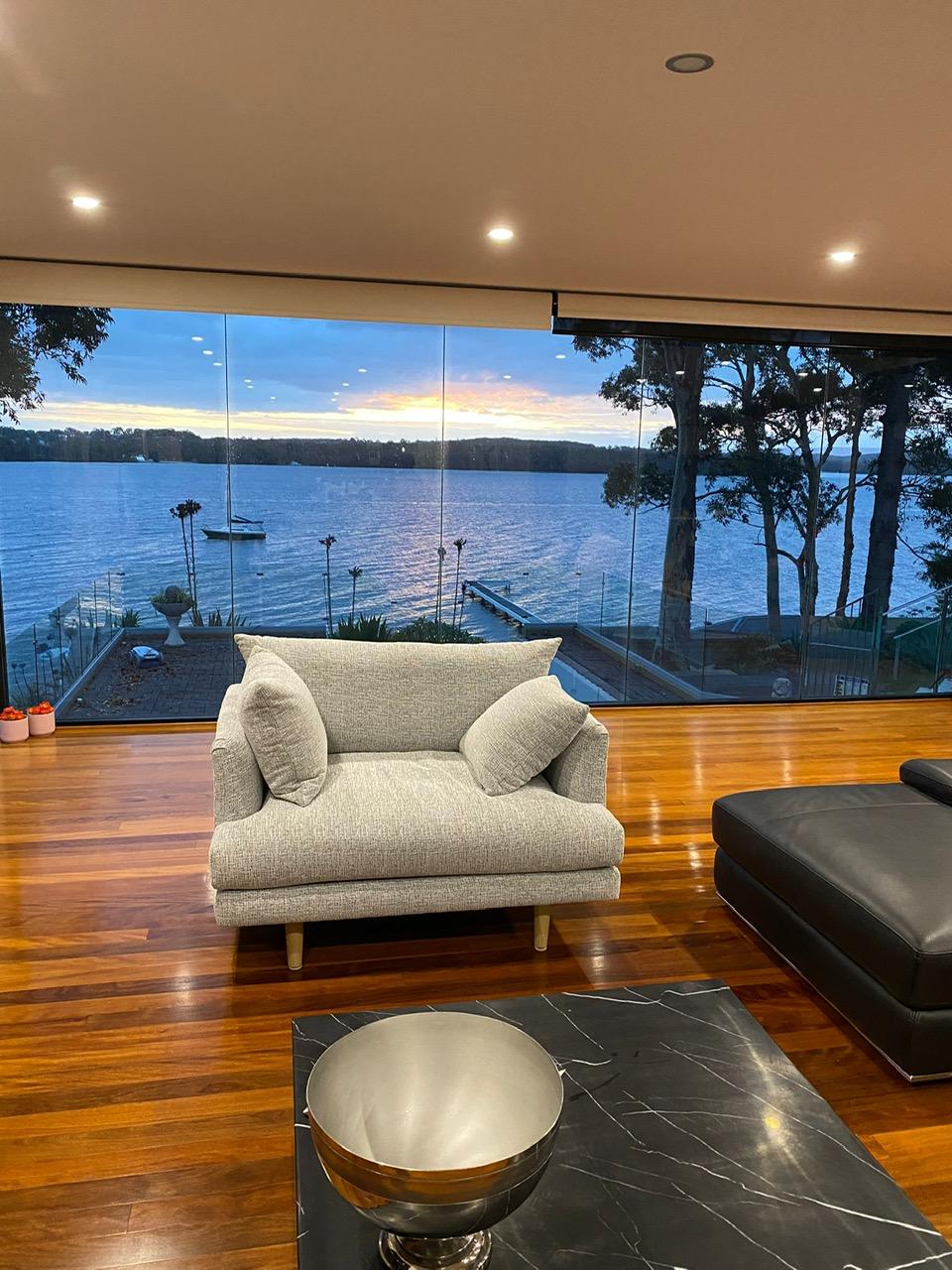 A cozy seating area is positioned next to expansive glass windows, showcasing stunning views of the waterfront at sunset. The floor features polished wooden planks, while a black coffee table sits within the space, complementing the elegant furnishings.