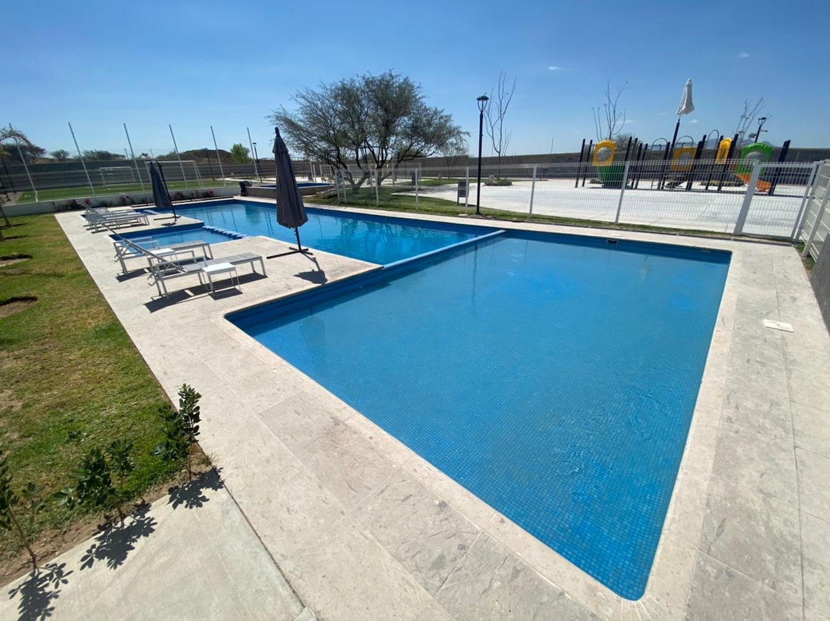 Two swimming pools are prominently featured, one for relaxation and the other for play. Lounge chairs are positioned around the pools, and a shaded umbrella stands near the water. In the background, a playground and green landscaping enhance the outdoor setting.