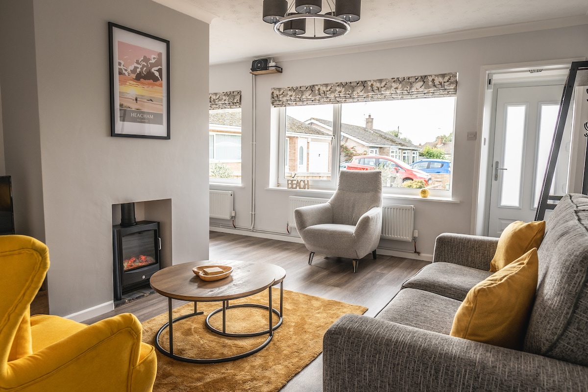 A bright lounge area features a comfortable gray sofa and a stylish yellow armchair. A round coffee table sits at the center, with an electric fireplace providing warmth. Large windows allow natural light to fill the space, with decorative shades offering privacy.