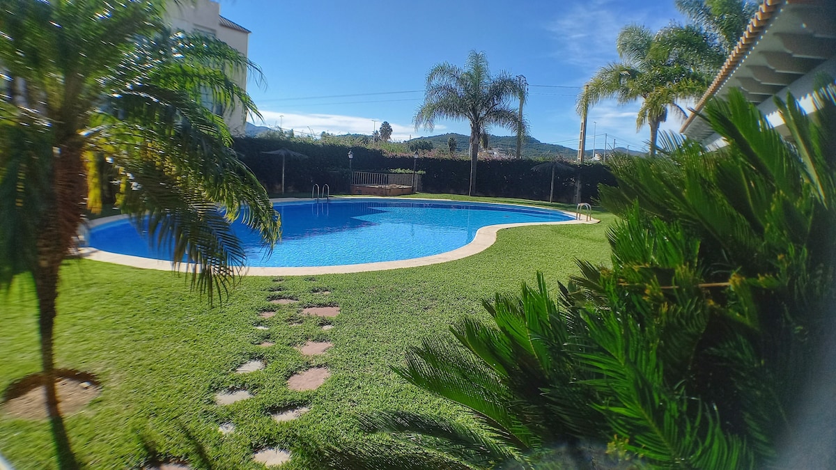 A large outdoor swimming pool is surrounded by manicured grass and palm trees. Sunlight reflects off the water, creating a serene setting. Steps lead into the pool, while a fence provides privacy from the surrounding area.