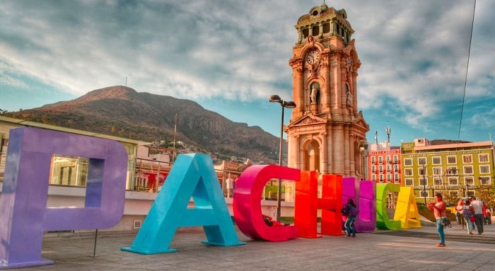 A vibrant display of oversized letters spelling 'PACHUCA' stands in a public space, with a historic clock tower visible in the background. The presence of a mountain adds depth to the landscape, while a gathering of people can be seen enjoying the area.