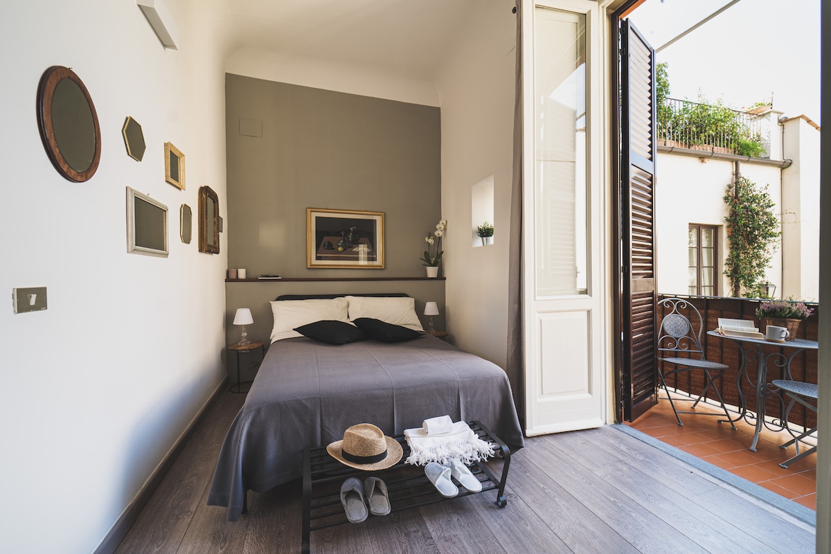 A spacious bedroom features a comfortable bed with neutral bedding. A large window opens to a small terrace with outdoor seating. Decorative mirrors and a framed artwork enhance the wall, creating a serene and functional environment.