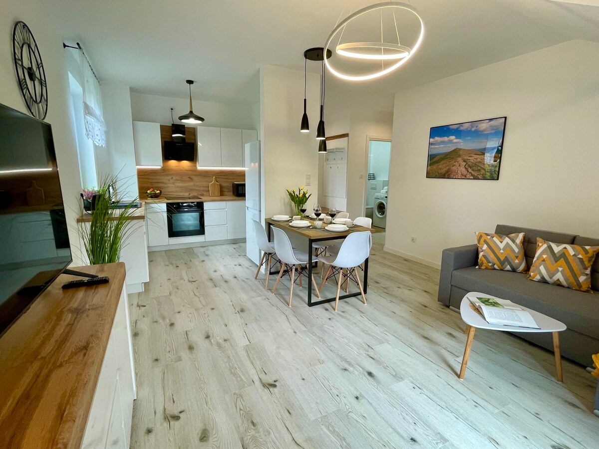 A modern living area is highlighted by a round dining table set for four. A sleek kitchen with minimalist cabinetry and a built-in oven is visible. Soft lighting fixtures hang above, and a comfortable couch with patterned cushions faces a television, creating an inviting space.