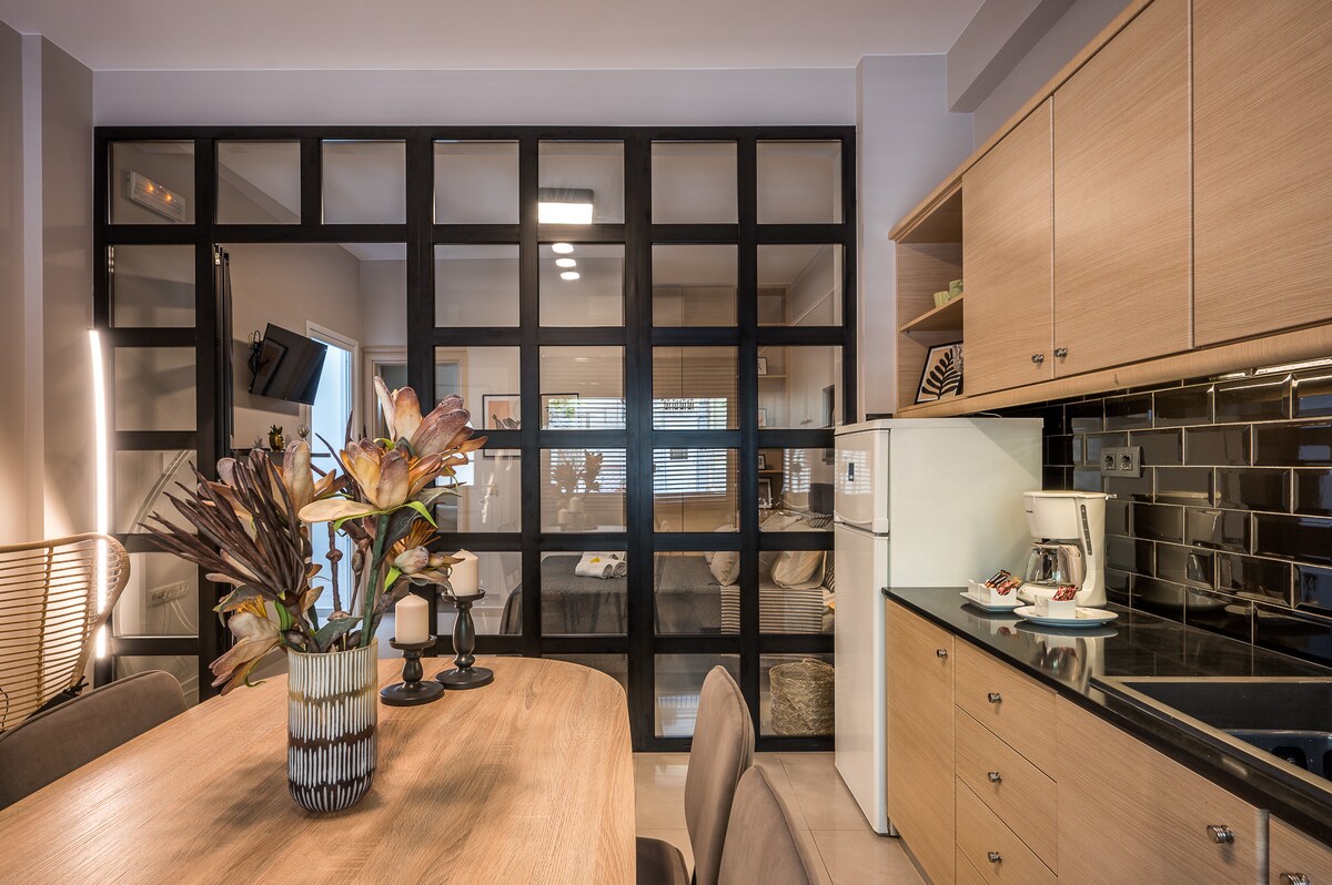 A modern kitchen area is featured with wooden cabinetry and a sleek countertop. A dining table with upholstered chairs is set, accompanied by a vase of flowers. Glass partition walls create an open yet defined living space, enhancing natural light throughout the area.