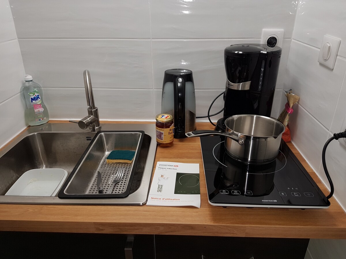 A compact kitchen space is presented, featuring a stainless steel sink with a dish rack. An electric kettle and coffee maker are positioned on the countertop, alongside an induction cooktop with a pot. Cleaning supplies and a user manual are also visible.