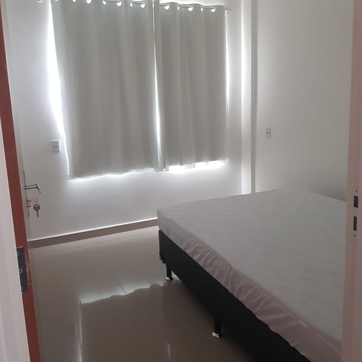 A simple bedroom is featured with light-colored walls and large windows covered by sheer curtains. A spacious bed without bedding is positioned centrally, and the glossy floor reflects the room's natural light.