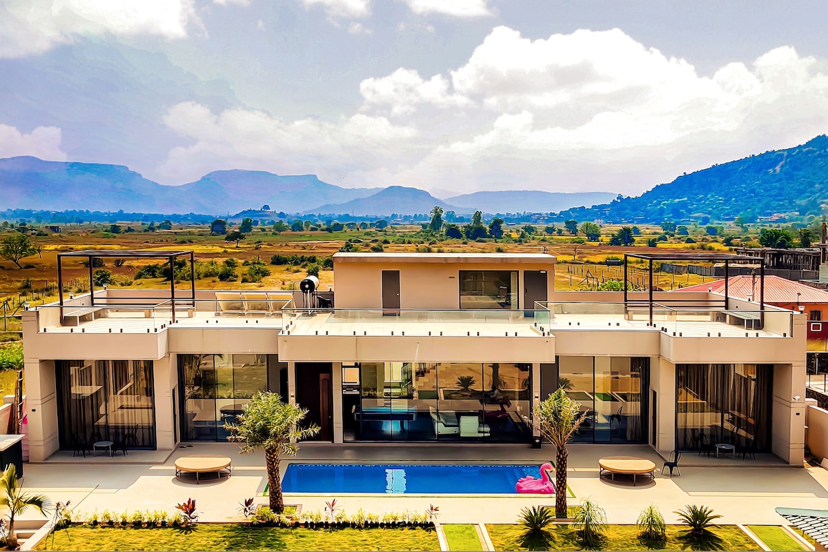 A modern villa is showcased with large glass windows reflecting views of surrounding hills. The outdoor area features a pool bordered by palm trees and loungers, creating a spacious layout. A rooftop terrace is visible, providing additional outdoor space for relaxation.