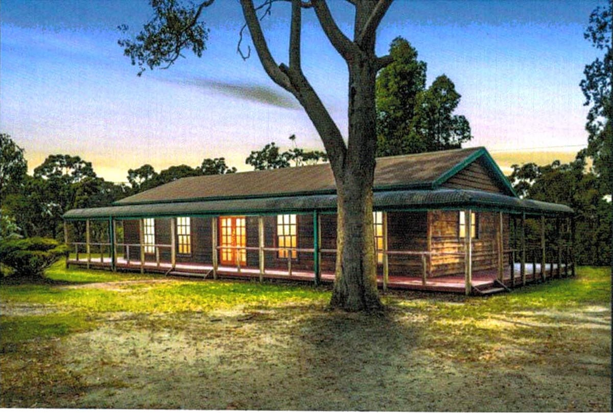 A spacious country-style home is surrounded by lush greenery, featuring a broad porch that wraps around the exterior. The windows glow softly with warm light, providing a contrasting appearance against the darkening sky.