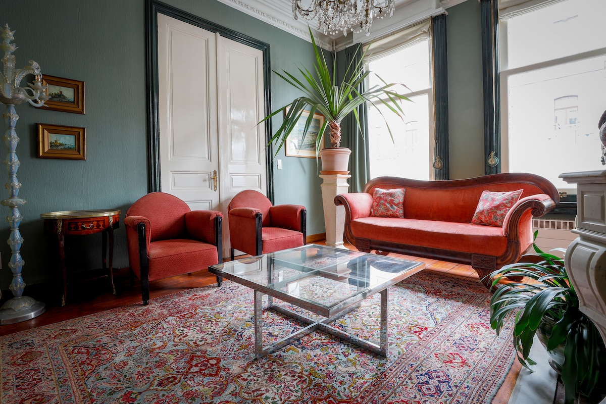 A spacious living area features two red armchairs and an elegant red sofa, positioned around a glass coffee table. An ornate chandelier hangs from the ceiling, while a large plant adds a touch of greenery. Natural light enters through two windows, illuminating the space.