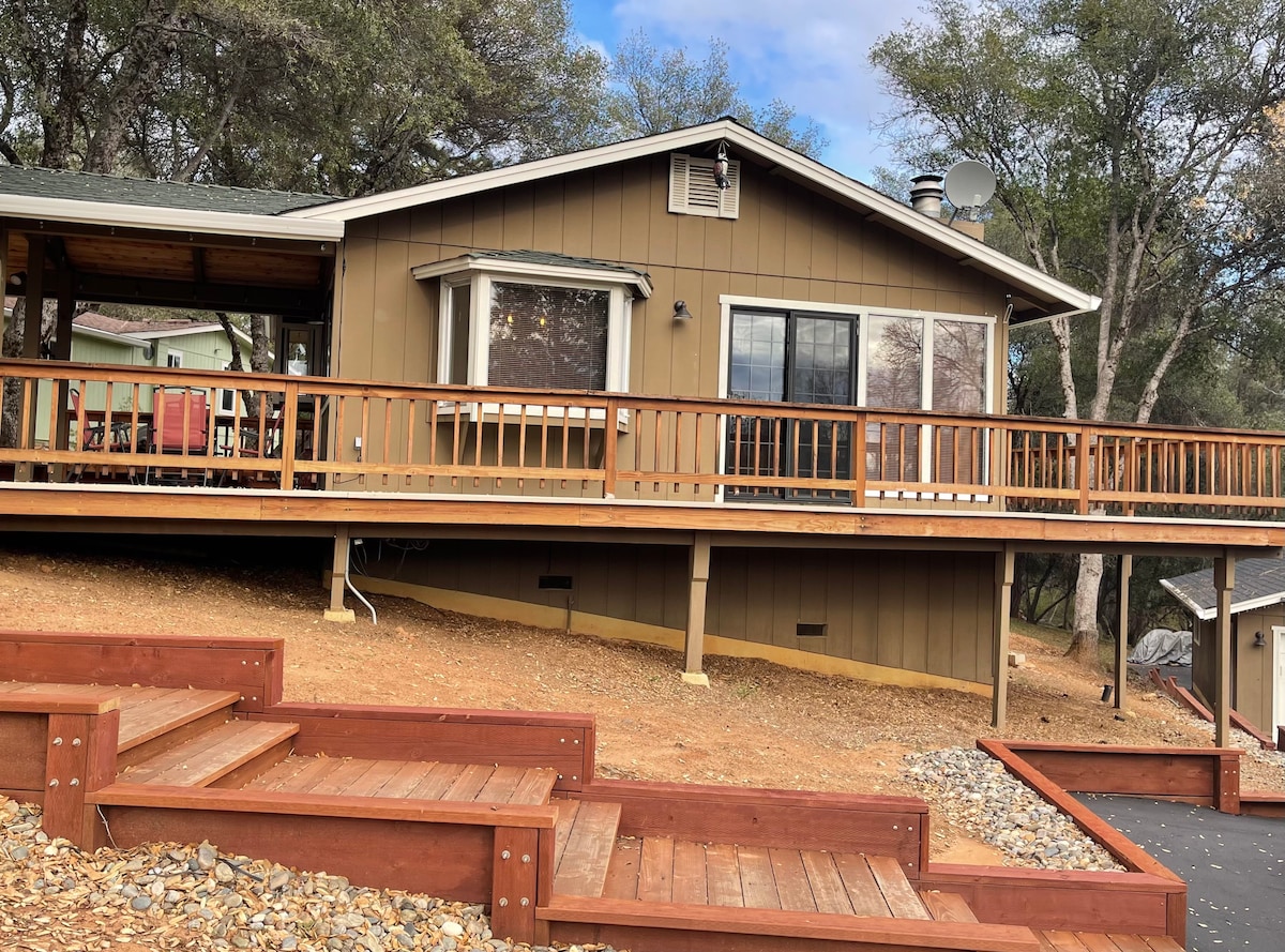 The exterior of the cabin features a large wrap-around deck with wooden railings, complemented by several windows providing ample natural light. The structure is set against a backdrop of trees and a blue sky, with steps leading up from the gravel path to the entrance.