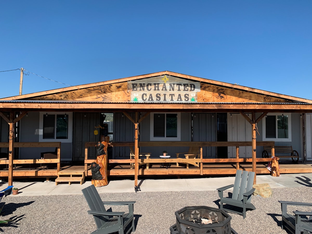 A welcoming facade of the Enchanted Casitas is presented, featuring wooden beams and a sign above the entrance. The porch is equipped with multiple seating options, and an arrangement of fire pit chairs is visible on the gravel surface, promoting outdoor relaxation.