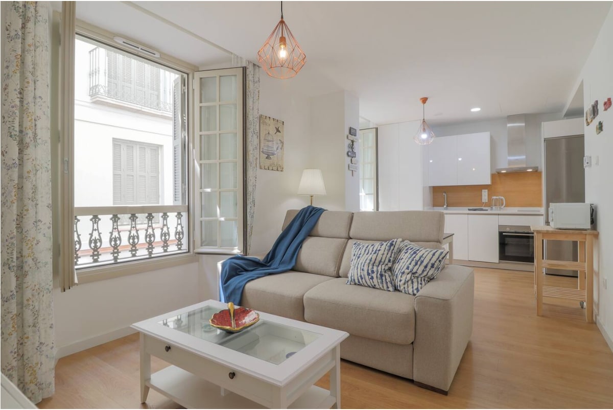 A light-filled living area features a comfortable sofa adorned with decorative pillows. Large windows offer views of the street and allow natural light to illuminate the space. A dining table is present, complemented by the modern kitchen in the background.