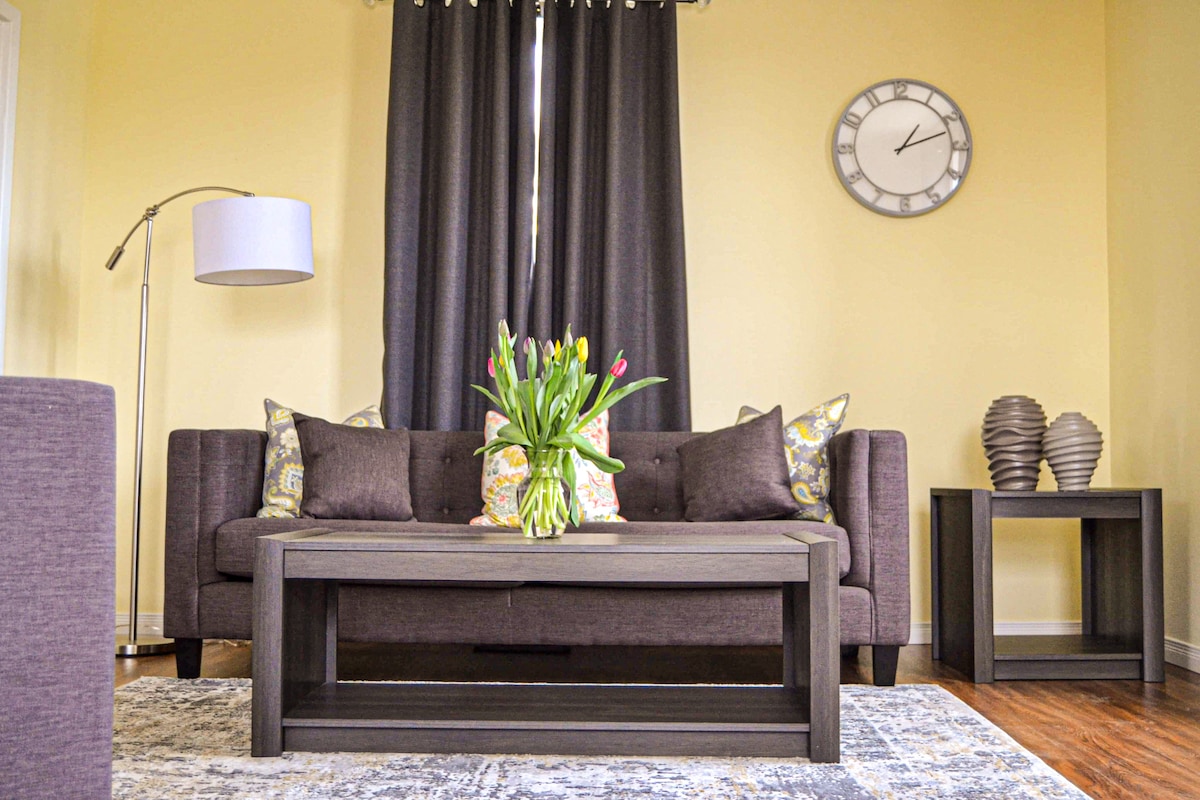 The living room features a dark sofa adorned with decorative pillows, complemented by a coffee table holding a vase of colorful tulips. A lamp stands beside the sofa, and a wall clock is displayed above. Warm yellow walls enhance the cozy ambiance.