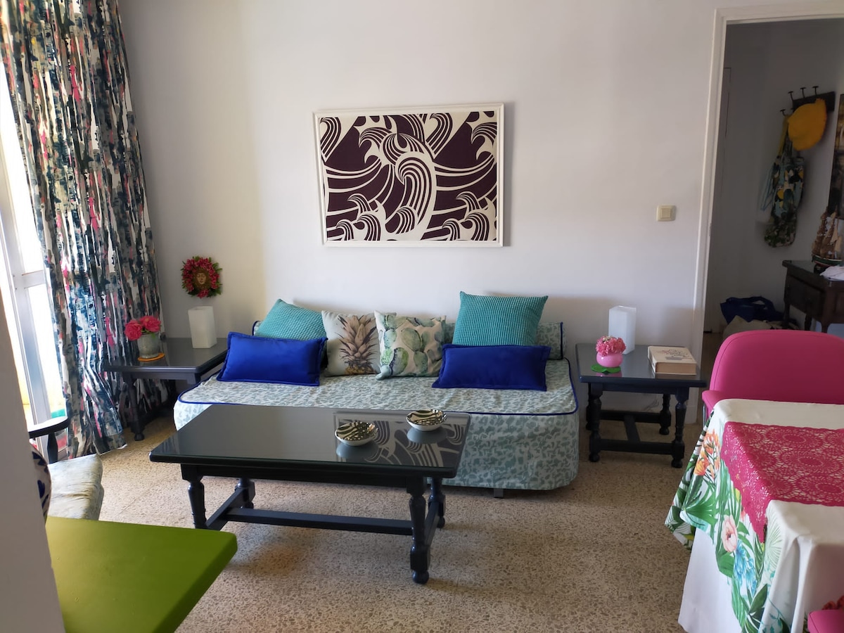 A bright living area features a patterned artwork above a comfortable sofa adorned with vibrant cushions. A dark wooden coffee table sits in front, complemented by side tables with decorative accents. Natural light is filtered through floor-to-ceiling curtains, casting a soft glow.