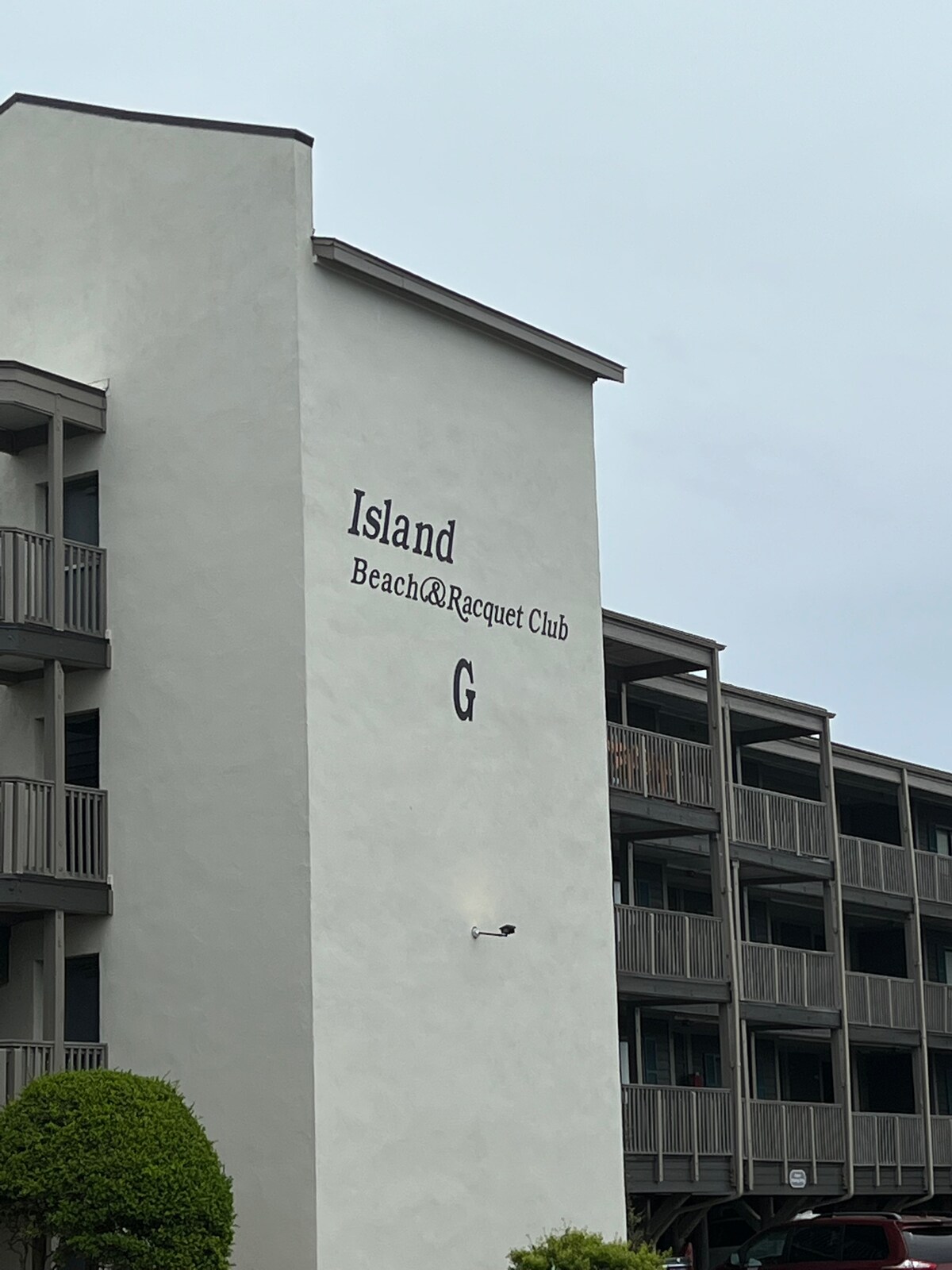 The building's exterior features a tall, white wall with the words 'Island Beach & Racquet Club' prominently displayed. Balconies line the structure, with several doors visible. A cloudy sky serves as the backdrop.